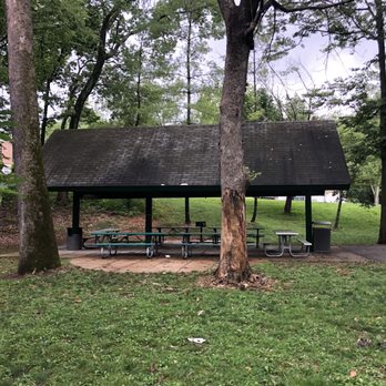 Stacy Park - A park shelter with a dark, sloped roof and open sides is situated on a paved area within a grassy park. Several picnic tables are arranged underneath the shelter, and a trash can is nearby. Tall trees surround the shelter, with some trunks showing signs of bark damage. The grass in the foreground is green with small patches of dirt and scattered leaves, indicating a natural outdoor setting. - Parks Near Me - Olivette, Missouri