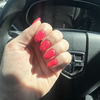 Sole Serenity - A close-up of a person's left hand displaying pink, glittery manicured nails is held up inside a vehicle. The background includes a steering wheel with a Dodge emblem and part of the vehicle's dashboard. The nails are well-groomed and painted with a bright pink polish containing glitter. The vehicle appears to be parked, with sunlight illuminating the scene. - Nail Salons Near Me - Tempe, Arizona