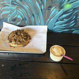 Vegan Gyro And The Vegan Ice Cream - A vegan gyro with a crumbly topping is placed on a metal tray lined with white paper, positioned on a dark wooden table. Next to it, there is a small cup of ice cream with a pink spoon inserted. The background features a wall with blue and teal abstract artwork and an electrical outlet.