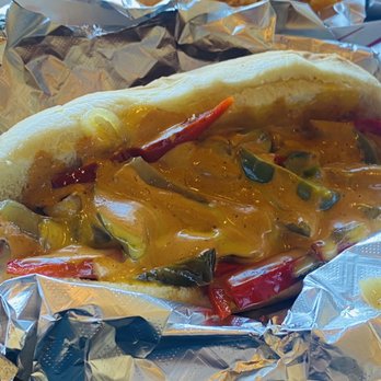 Dogos VIP - Philly cheesesteak dog - A hot dog in a soft bun is topped with melted cheese, sliced red and green peppers, and sauce. The hot dog is partially wrapped in crinkled aluminum foil, placed on a surface with other foil wrappers visible in the background. - Hot Dogs Near Me - Albuquerque, New Mexico