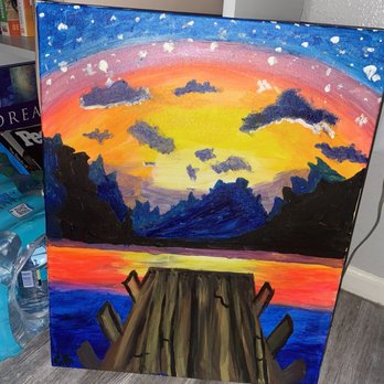 Pinot's Palette - The painting shows a sunset scene with vibrant colors, including orange, yellow, and pink, over a mountain range and water. A wooden dock extends from the bottom center into the water, with visible cracks and uneven planks. Dark clouds are scattered across the sky, which transitions from starry night to sunset hues. The scene captures a tranquil, natural landscape with a focus on the dock as the foreground element. - Paint & Sip Near Me - Gilbert, Arizona