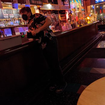 Big Chicks - A person in a tropical-patterned shirt and mask is leaning on a bar counter in a lounge with colorful artwork and neon signs. - Gay Bars Near Me - Chicago, Illinois