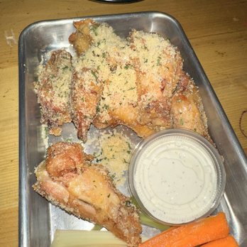 Eli Cannon's - A metal tray contains breaded chicken wings garnished with herbs, accompanied by a small container of creamy dipping sauce. There are also celery sticks and carrot sticks placed on the tray. The tray is on a wooden table, with part of a black object visible in the background. - American Near Me - Middletown, Connecticut