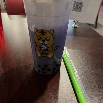 Tasty Pot - Taro milk tea boba - A plastic cup of bubble tea with a cartoon tiger holding a drink printed on it, placed on a dark wooden table. The cup has a clear plastic lid, and a green straw is lying next to it in a plastic wrapper. In the background, there is a sign with a QR code and a red container, indicating a casual dining or cafe setting. - Hot Pot Near Me - Albuquerque, New Mexico