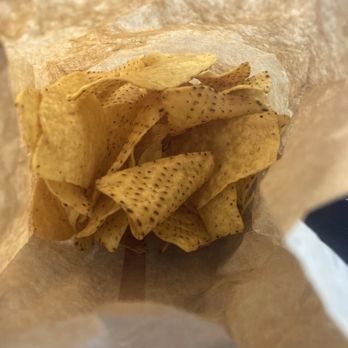 Gracie's Burritos - CHIPS AND SALSA - The image shows the interior of a paper bag containing multiple tortilla chips. The chips are triangular, light yellow, and have black specks, indicating seasoning or spices. The bag's interior is plain, and the chips are piled loosely, filling the bottom of the bag. - Mexican Near Me - Monee, Illinois