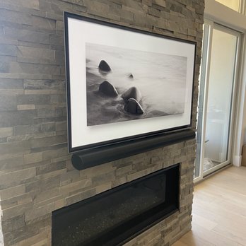 Marvel Audio - Tv and sound bar installation - A black-and-white photograph depicting rocks on a beach is mounted in a black frame with a white mat. The photograph is positioned above a modern, rectangular fireplace with a black trim, set into a textured stone wall. To the right, there is a sliding glass door allowing natural light into the space, with a wooden floor beneath. - Home Theatre Installation Near Me - Folsom, California