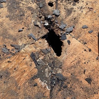 First Response Roofing - Before First Response - The image shows a wooden surface with a significant hole in the center, surrounded by splintered wood and debris. The surface appears weathered and damaged, with scattered fragments of wood and dark material around the hole, indicating possible structural damage or impact. - Roofing Near Me - Gilbert, Arizona