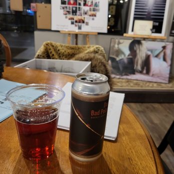 Black Dog Vinyl Cafe - A can of beer labeled 'Dark Side of the Bad Pole' and a plastic cup filled with a dark beverage are placed on a wooden table inside a cafe. The background features framed artwork, photographs, and a window showing the outside night scene. The setting appears casual with additional items like a menu and a fur-lined coat or blanket on a chair. - Coffee & Tea Near Me - Plainfield, Illinois