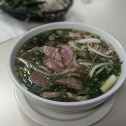 Combination pho - A bowl of Vietnamese pho contains sliced beef, chopped green onions, cilantro, and bean sprouts in clear broth. The bowl is placed on a white table, with a basket of fresh greens and herbs in the background. The image captures the dish's ingredients and presentation typical of Vietnamese cuisine.