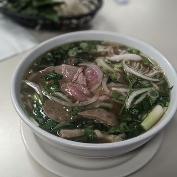 Pho Leo - Combination pho - A bowl of Vietnamese pho contains sliced beef, chopped green onions, cilantro, and bean sprouts in clear broth. The bowl is placed on a white table, with a basket of fresh greens and herbs in the background. The image captures the dish's ingredients and presentation typical of Vietnamese cuisine. - Vietnamese Near Me - Mesa, Arizona