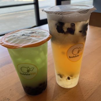 Bubblebee X Ramen - Green Apple Green Tea and Blueberry Lychee Fruit Tea - Two bubble tea drinks with tapioca pearls are positioned on a wooden table inside a cafe. The drink on the left is in a green cup with an orange lid, while the drink on the right is in a clear cup with a plastic lid. The background features a large window revealing a parking lot with cars and some greenery outside. The drinks are labeled with the Bubble Bee logo. - Ramen Near Me - Ashburn, Virginia