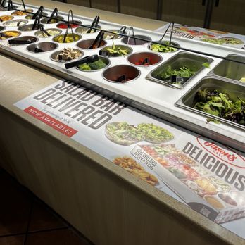 Jason's Deli - A salad bar with multiple containers holding fresh vegetables, toppings, and dressings, with a promotional sign on the counter. - Delis Near Me - Albuquerque, New Mexico