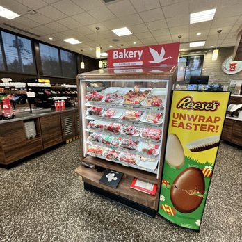 Wawa - A bakery display case containing various packaged baked goods is positioned inside a store, with a sign above indicating 'Bakery' and 'Fresh Baked Daily.' To the right, a colorful Reese's Easter promotional sign features images of chocolate eggs and candies. The store interior includes ceiling lights, large windows, and a counter area with various items in the background. - Gas Stations Near Me - Maple Shade, New Jersey