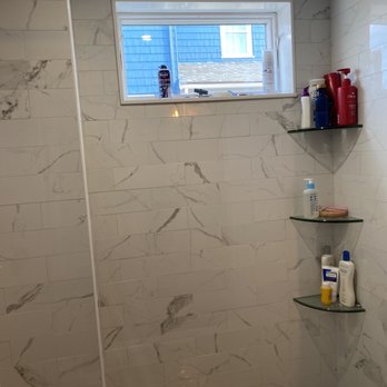 J.F. Murray Construction - 1st floor shower - The image shows a shower area with walls covered in marble-patterned tiles. There are three glass corner shelves mounted on the right wall, holding bottles of shampoo, conditioner, and other toiletries. A small window near the ceiling provides natural light, with a view of neighboring houses and a blue sky. The ceiling appears to be tiled, and the shower floor is visible at the bottom of the image. - General Contractors Near Me - Stoneham, Massachusetts