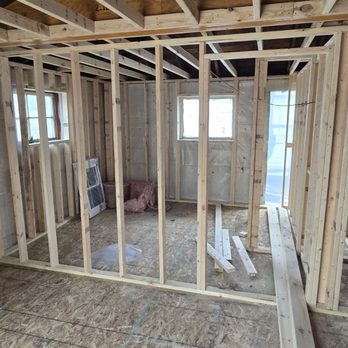 Morpho Carpentry - The photo depicts the interior of a building under construction, featuring exposed wooden framing including vertical studs, horizontal beams, and roof trusses. Two small windows are installed, allowing natural light into the space. The floor is covered with plywood sheathing, and construction materials are scattered around. The framing suggests the early stages of building a room or house, with structural elements in place but no interior finishes yet. - General Contractors Near Me