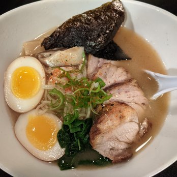 Ebisu - Miso Ramen - pork miso broth, flavored soft boiled egg, shiitake mushroom, bamboo shoots, spinach, seaweed, fish cake, spices, chashu - A bowl of Japanese ramen includes two halved soft boiled eggs with runny yolks, slices of pork, a piece of seaweed, chopped green onions, and broth. The dish is presented in a white bowl with a white spoon on the side, showcasing traditional ramen ingredients and garnishes. - Sushi Bars Near Me - Providence, Rhode Island