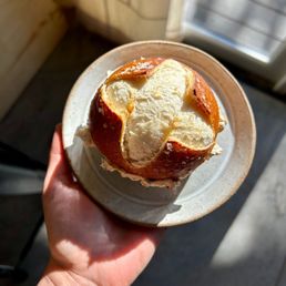 A person holds a small ceramic bowl with a partially eaten bread roll, set on a small plate, near a window with sunlight.