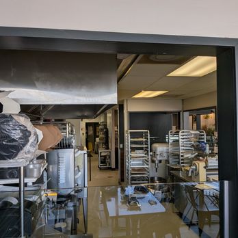 Sucre Table - The image shows the interior of a bakery kitchen viewed through a large window. Inside, there are metal baking racks, a stand mixer, and various baking tools and supplies. The space appears organized with equipment and ingredients arranged for baking operations. The lighting is bright, and the overall setting suggests a professional bakery environment. - Bakeries Near Me - Tampa, Florida