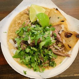 The image shows a taco with a corn tortilla base topped with shredded meat, chopped cilantro, and diced onions. Two lime wedges and grilled pineapple slices are placed on top, garnishing the dish. The taco is served on a white plate with a piece of parchment paper underneath, placed on a wooden surface. The presentation highlights traditional Mexican ingredients and flavors.