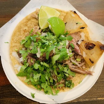 Taco Libre - The image shows a taco with a corn tortilla base topped with shredded meat, chopped cilantro, and diced onions. Two lime wedges and grilled pineapple slices are placed on top, garnishing the dish. The taco is served on a white plate with a piece of parchment paper underneath, placed on a wooden surface. The presentation highlights traditional Mexican ingredients and flavors. - Mexican Near Me - Minneapolis, Minnesota