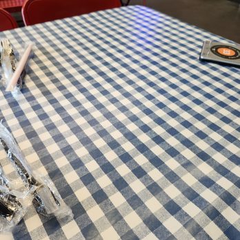 Mighty Fine Burgers Cedar Park - Tablecloth - The image shows a table covered with a blue and white checkered tablecloth. On the table, there are wrapped utensils and a menu or promotional card. Red chairs are positioned behind the table, and part of a checkered cloth is visible in the background. The setting appears to be a casual dining area, possibly in a restaurant or cafe. - Burgers Near Me - Cedar Park, Texas
