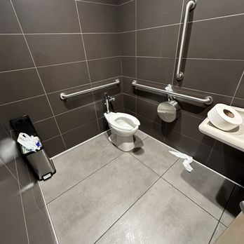 McDonald's - yucky bathroom - The image shows a public restroom with gray tiled walls and floor. A toilet is positioned against the wall, equipped with metal grab bars on both sides for accessibility. A trash bin is placed in the corner, and a paper roll dispenser is mounted on the wall. There is some trash on the floor near the toilet. The restroom appears clean but has some minor disarray with tissue paper on the floor. - Fast Food Near Me - Independence, Ohio