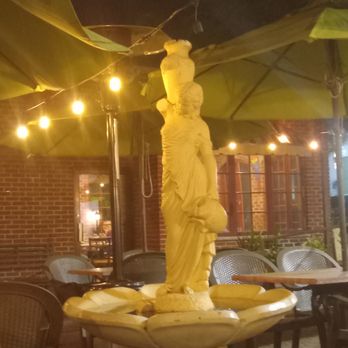 La Strada Restaurant - The outside area. - The image shows a stone statue of a woman holding a child, positioned in the center of an outdoor restaurant seating area. The area features tables with chairs, large green umbrellas, and string lights providing warm illumination. The background includes brick walls and windows, indicating a cozy, outdoor dining space likely associated with a restaurant or cafe. - Pizza Near Me - Millburn, New Jersey