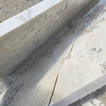 Benchmark Surfaces - Mishandled my remnants and cracked it, and did not let anyone know before hand, just left the pieces outside. - The image shows a marble or stone slab with a visible crack running through its surface, placed on a concrete ground. The slab appears to be part of a construction or renovation project, with a textured surface and natural veining. A mesh or protective covering is partially visible at the top of the image, and a shoe is seen in the background, indicating an outdoor setting. - Countertop Installation Near Me