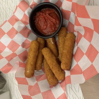 Stanley's Restaurant - Mozzarella sticks - Six mozzarella sticks with a golden-brown breaded coating are arranged on red and white checkered paper. A small black cup filled with marinara sauce is positioned at the top center of the basket. The basket is placed on a white surface with a subtle pattern. The presentation suggests a casual appetizer or snack, typical of American comfort food. - Seafood Near Me - Central Falls, Rhode Island