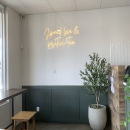Interior of a tea shop featuring a white wall with a neon sign that says 'Spread love & positive tea,' a potted plant, and wooden furniture.