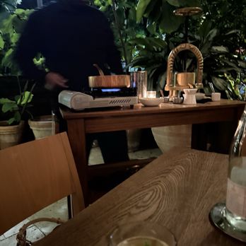 Ariete - Duck - A person is preparing food outdoors at a wooden table with a portable stove, surrounded by lush green plants and potted foliage. - New American Near Me - Miami, Florida