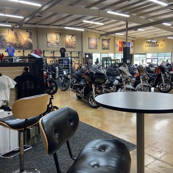 Willamette Valley Harley-Davidson - The image depicts the interior of Willamette Valley Harley-Davidson, a licensed motorcycle dealership in Eugene, OR. The showroom displays multiple Harley-Davidson motorcycles arranged in rows near large windows. Merchandise such as T-shirts and helmets are visible on racks and shelves along the walls. The space includes seating with chairs and a high table, and promotional photographs of motorcycles are hung on the walls. The dealership serves the Willamette Valley region, including Portland and Eugene. - Motorcycle Dealers Near Me - Eugene, Oregon