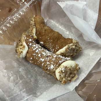 Sergio's Bakery and Cafe - Cannolis $2.50 each - A cannoli pastry with a fried, crispy shell filled with a creamy, white filling, topped with a light dusting of powdered sugar. The pastry is partially dipped in chopped nuts at the ends. It is placed inside a clear plastic container lined with parchment paper, on a wooden surface. - Bakeries Near Me - Albuquerque, New Mexico