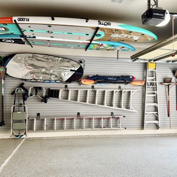 Classy Closets - Garage wall features organized sports equipment and tools, including surfboards mounted on the ceiling, paddles, a ladder, and gardening tools such as a rake and shovel, all arranged on a slatwall panel for storage and accessibility. - Cabinetry Near Me - Grapevine, Texas