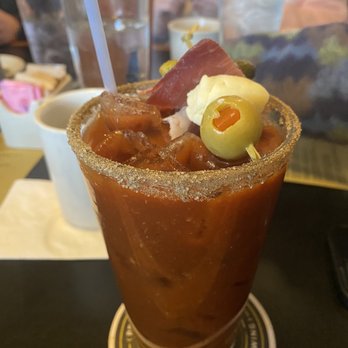 The Sample Room - Housemade Zesty Bloody Mary - A tall glass contains a Bloody Mary cocktail with a salted rim, filled with a tomato-based drink and ice. The garnish includes green olives, a pickle, and a slice of meat, with a straw inserted. The background shows other drinks and table items, indicating a restaurant or bar setting. - New American Near Me - Minneapolis, Minnesota