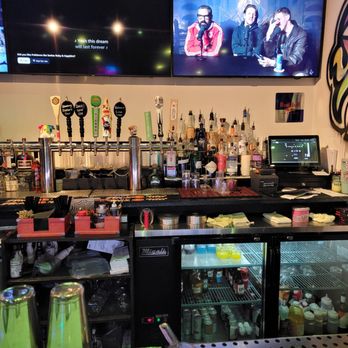 Three Lives - A bar counter with multiple beer taps labeled with different flavors, bottles of various spirits, and a refrigerated display case containing bottled drinks. Two large screens are mounted above the bar, displaying content and a podcast recording with three individuals. The setup includes bar tools, napkins, and other bar accessories, indicating a lively, casual environment at Three Lives in Syracuse, NY. - Burgers Near Me - Syracuse, New York