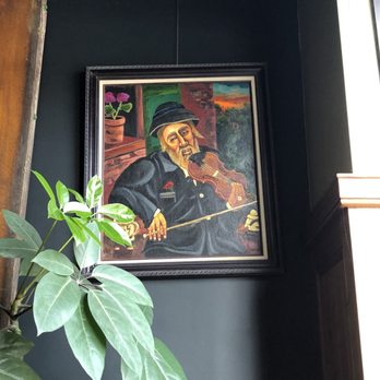 Bar La Tosca - Interior - A painting of an elderly man with a beard, wearing a hat and dark clothing, playing the violin, is displayed on a dark wall near a window. - Italian Near Me - Ames, Iowa