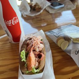 Lobster roll - A lobster roll with lettuce and lobster meat in a paper tray is placed on a wooden table. Nearby, there is a Coca-Cola cup, a bag of potato chips, and some food wrappers. In the background, there are additional food items, a smartphone, and a person wearing a gray jacket. The setting appears to be a casual dining environment, possibly at a seafood restaurant or food stand.
