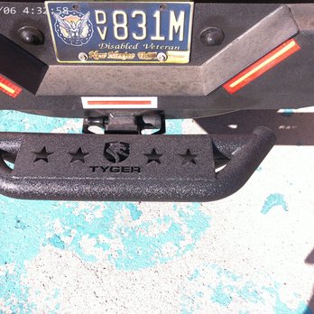 Auto Depot - The photo captures the rear section of a vehicle, featuring a New Mexico disabled veteran license plate with the number V831M. Below the license plate, there is a TYGER brand bumper guard with star-shaped cutouts. The vehicle is parked on a surface with peeling blue paint, and the image includes a timestamp in the top left corner. The shadow of the vehicle is visible on the ground. - Auto Detailing Near Me - Albuquerque, New Mexico
