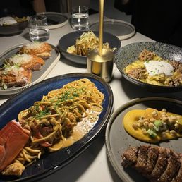 Seared Duck Breast - The image shows a dining table set with several plated dishes including a pasta dish with green herbs, a steak with sauce, and other meals with garnishes. There are glasses of water and a modern, gold-colored lamp providing lighting. The setting appears to be in a restaurant with a dark background and a sophisticated ambiance.