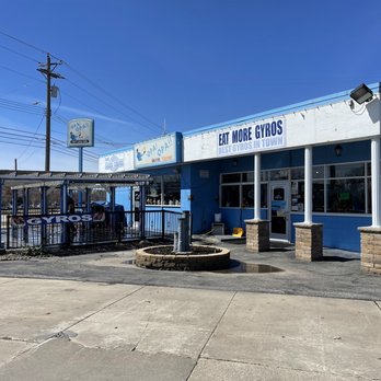 Opa! Opa!! - a blue building with a blue awning - A Greek restaurant painted in blue with signage that promotes gyros and other Greek dishes. The building has large windows and a covered outdoor seating area enclosed by a metal fence, with a banner advertising gyros. The parking lot and sidewalk are visible in the foreground, and utility poles and wires are seen in the background. The sky is clear and blue. - Greek Near Me - Rochester, Minnesota