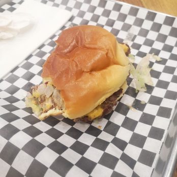 Simple Burger - Simple burger - A partially eaten burger featuring a soft bun, beef patty, melted cheese, and sliced onions is presented on a checkered paper-lined tray. The tray is on a wooden table, with some lettuce and crumbs scattered around. The burger appears to be served in a casual dining setting. - Burgers Near Me - Toronto, Ontario