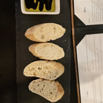 Debajo Tapas ABQ - Four slices of bread with a light, airy texture and irregular holes are placed on a black rectangular serving plate. A small square dish containing olive oil with balsamic vinegar drizzled on top is positioned at the top of the plate. The background includes a partially visible menu with the word 'Wine' and some text, indicating a dining setting. - Spanish Near Me - Albuquerque, New Mexico