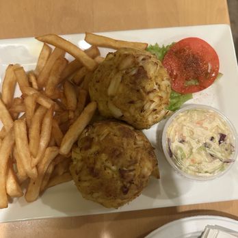 Koco's Pub - 2 crab cake platter - A square white plate holds two crab cakes, French fries, a slice of tomato on lettuce, and a small cup of coleslaw. The crab cakes are round and golden-brown, with visible bits of crab meat. The French fries are crispy and piled on one side of the plate. A slice of tomato rests on a leaf of lettuce, and a small cup of coleslaw with shredded vegetables and dressing is included. The plate is placed on a wooden table. - Pubs Near Me - Baltimore, Maryland
