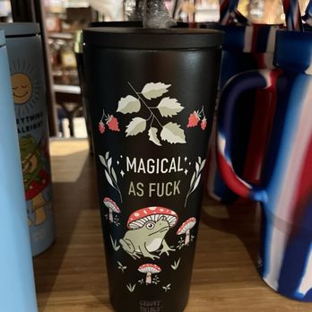 Atown - I loved this cup! - A black travel mug with a lid and straw, decorated with whimsical illustrations of mushrooms, a frog, and leaves. The mug has the phrase "Magical As F**k" printed prominently in the center. It is placed on a wooden surface among other colorful mugs and items, suggesting a retail setting that offers unique and playful products. - Accessories Near Me - Austin, Texas
