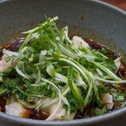 Spicy wonton appetizer - A ceramic bowl contains an Asian noodle dish with broth, topped with fresh green onions, cilantro, and microgreens. The dish appears to include dumplings or similar ingredients, with a dark soy-based sauce visible in the broth. The setting suggests a restaurant environment with a wooden table surface.