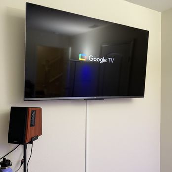 24K CCTV - Office TV mounted - A flat-screen Google TV is mounted on a white wall, with a visible white cable running down from it. A small speaker with a wood finish is mounted below the TV. - Security Systems Near Me