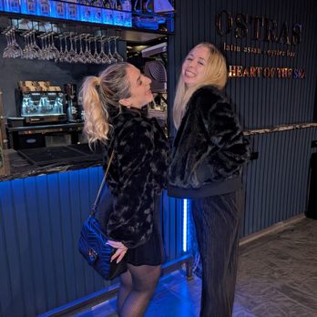Ostras - Two women wearing black fur coats are standing close together, smiling and laughing at each other inside Ostras, a Latin-Asian fusion oyster bar. One woman has a blonde ponytail and is holding a small black purse with a chain strap, while the other has long blonde hair. The background features a bar counter with glassware, bottles, and a coffee machine, along with a sign that reads 'Ostras Latin Asian Oyster Bar' and 'Heart of the Sea.' The setting has a dark, modern interior with blue lighting accents. - Asian Fusion Near Me - Des Plaines, Illinois