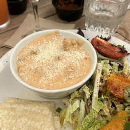 Wedding Chicken - The image shows a plate with a side salad, a bowl of creamy dip topped with cheese, and folded tortillas. A cocktail with foam and a coffee bean garnish is placed behind the plate, along with a glass of water. The setting appears to be a restaurant table with condiments and utensils visible in the background.