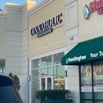Cinnaholic - The image shows the exterior of Cinnaholic, a bakery specializing in cinnamon rolls, with a prominent sign displaying the name 'Cinnaholic' and the tagline 'Gourmet Cinnamon Rolls.' The storefront features large glass windows and a glass door, with a green hedge in a rectangular planter in front. Adjacent to the bakery, there is a green Huntington Learning Center sign and window advertisements for tutoring services. The building has a beige facade with architectural details and a clear blue sky overhead. - Bakeries Near Me - Gambrills, Maryland